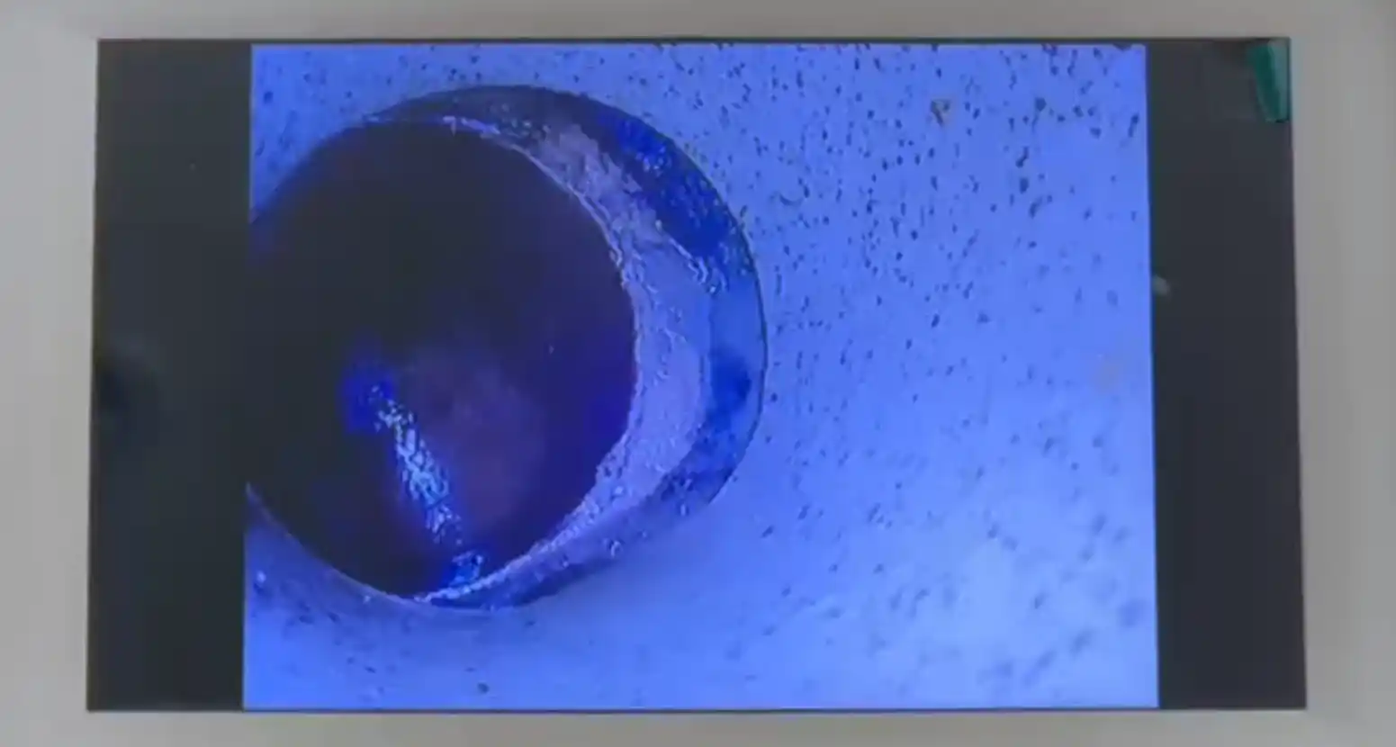 A high-definition CCTV camera view inside a residential drain pipe showing a circular opening with mineral buildup and a partial blockage along the inner wall.