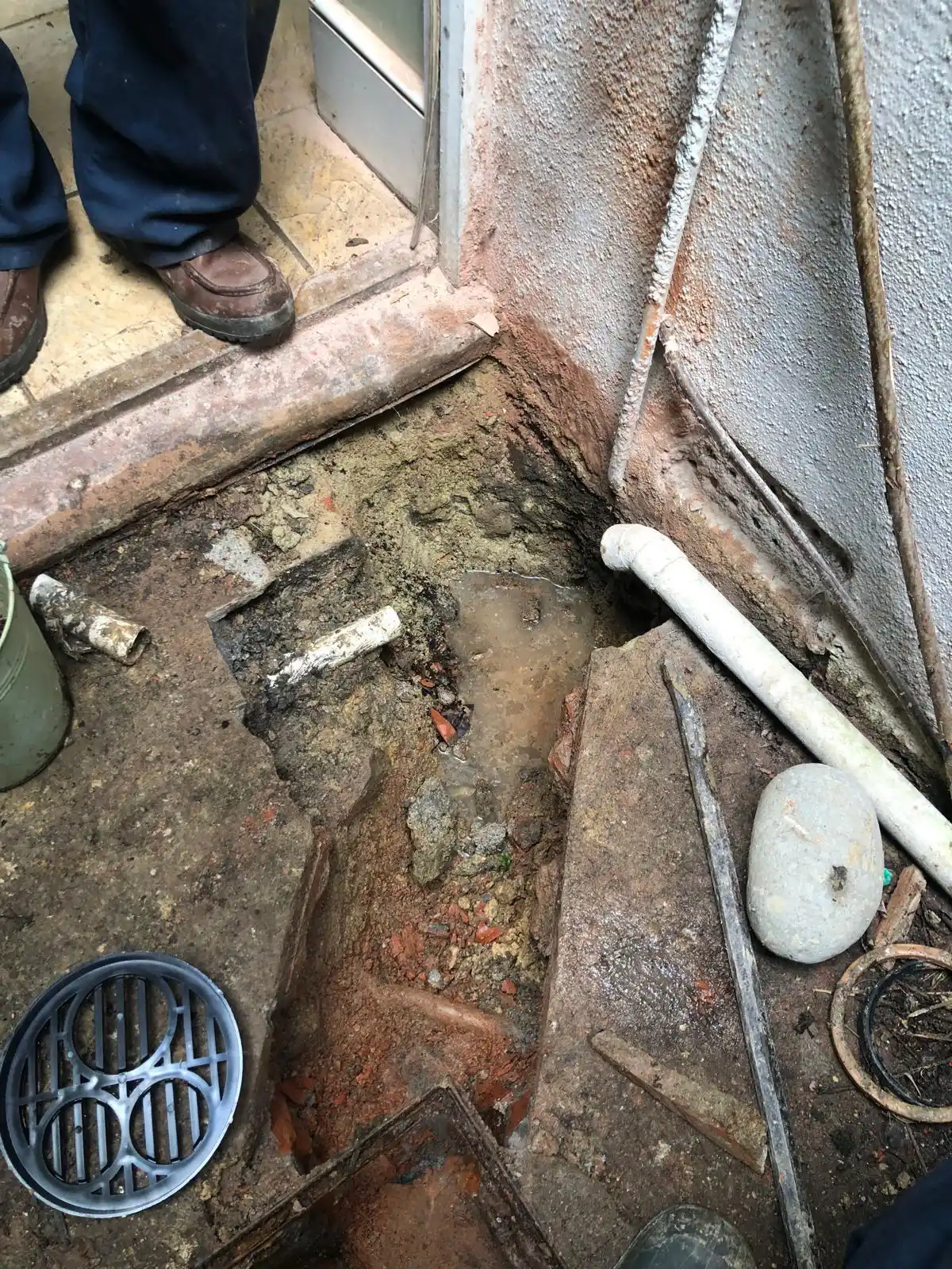 Open trench in a concrete floor beside a doorway exposing soil and a white PVC pipe, with a dislodged metal grate, scattered tools, and a worker’s boots visible during a drain repair.