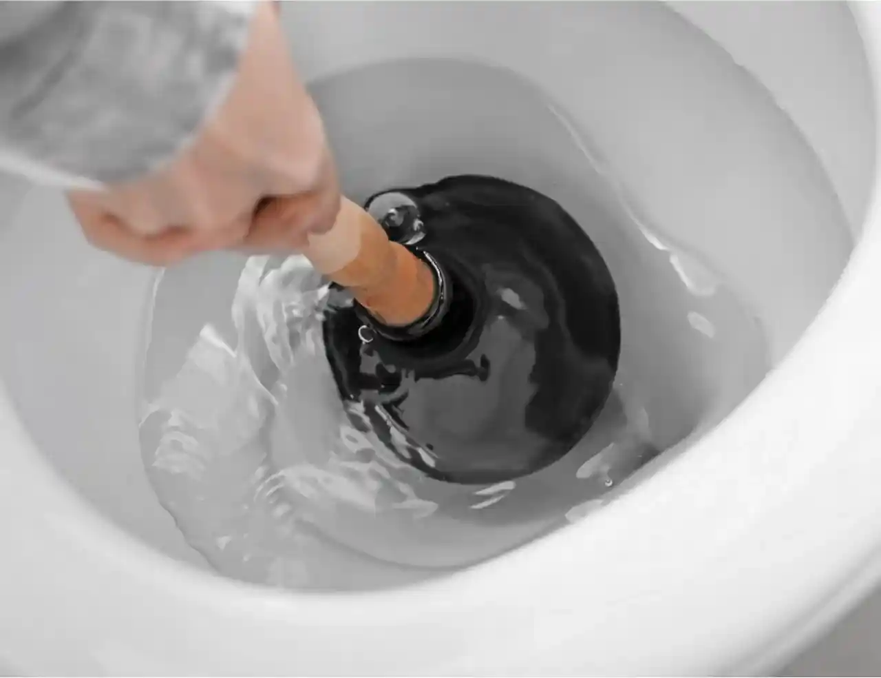 A hand grips a wooden-handled plunger, forcing a black rubber cup over the drain hole of a white porcelain toilet bowl filled with rippling water.