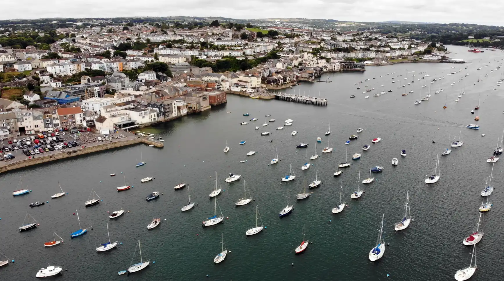 Aerial view of boats moored in the Penryn River, looking toward the town of Falmouth, Cornwall — with a dense cluster of coastal homes and harbour buildings.