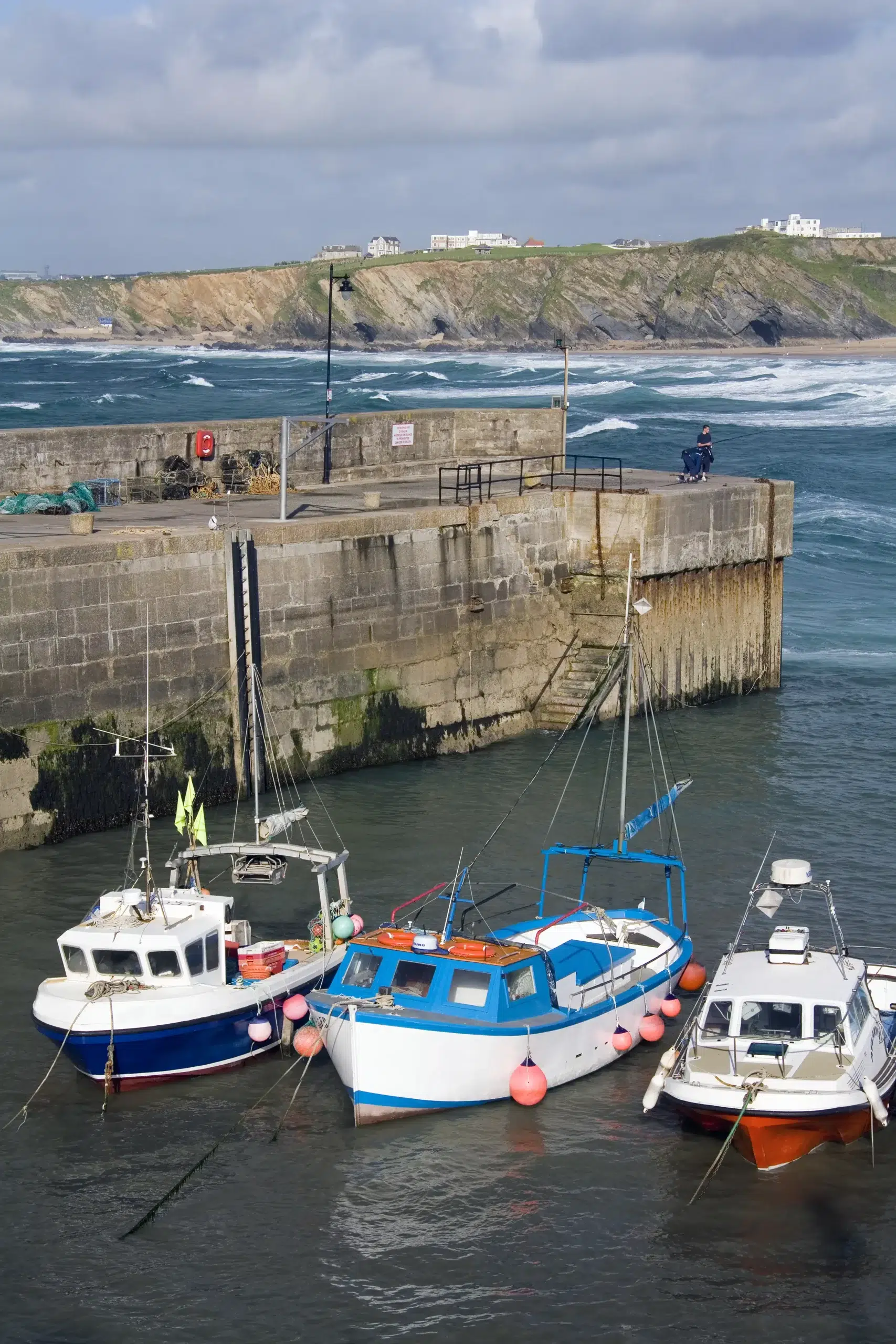 Fishing boats moored at Newquay Harbour in Cornwall, with a person seated on the quay and waves crashing against the cliffs in the background.