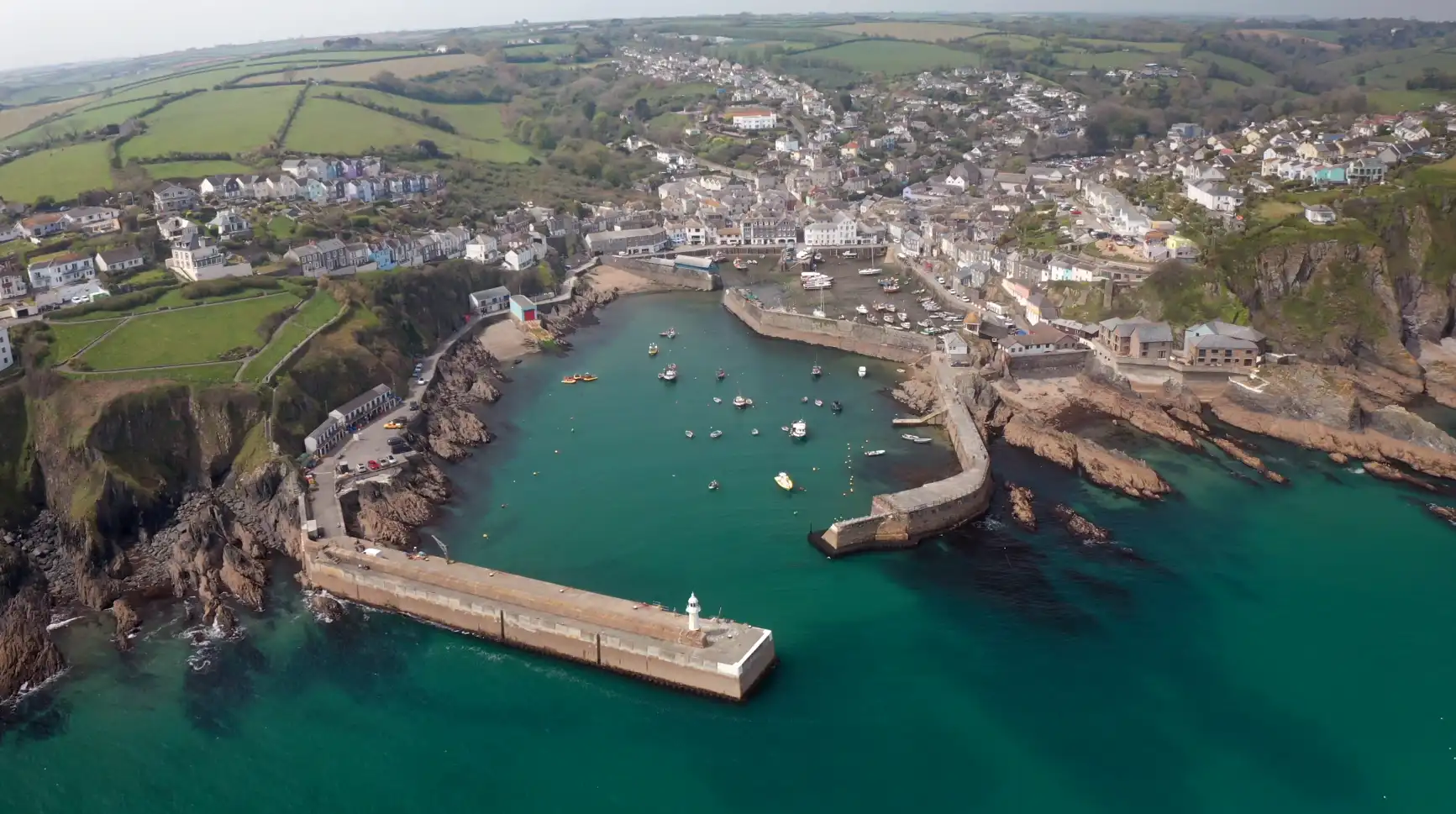 Aerial view of Mevagissey Harbour in Cornwall, showing colourful homes, fishing boats, and the surrounding rocky coastline and green fields.