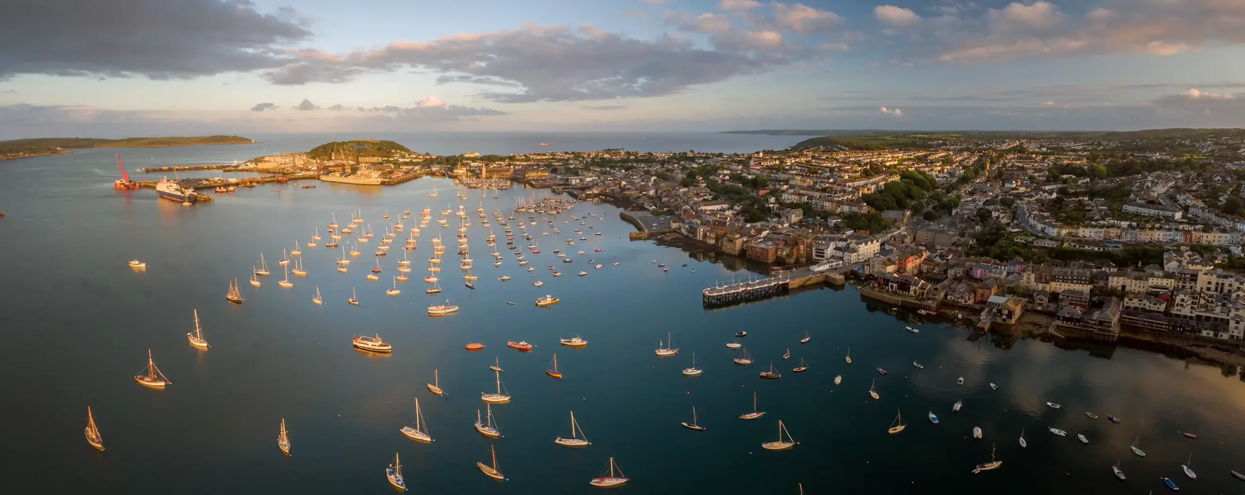 Panoramic aerial view of Falmouth harbour filled with boats at sunset — showing the coastline, town centre, and residential areas of West Cornwall.