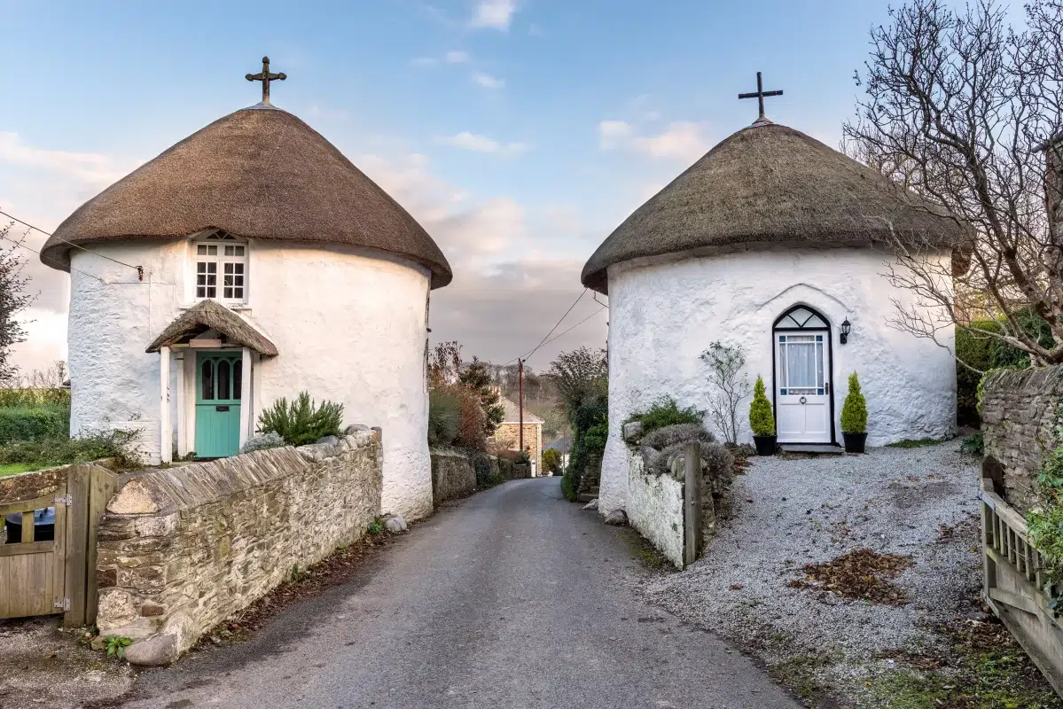 Historic roundhouses with thatched roofs in the village of Veryan, Cornwall — showcasing traditional architecture on a narrow rural lane.