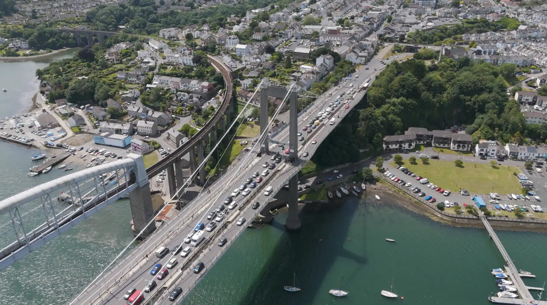 Aerial view of Tamar Bridge and Royal Albert Bridge spanning the River Tamar into hilly Saltash, crowded with traffic and surrounded by Victorian houses and boat moorings.