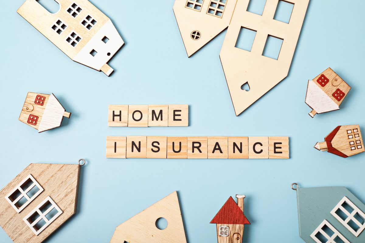 Flat-lay of small wooden house cut-outs encircling scrabble tiles that spell “HOME INSURANCE” on a pale-blue background, symbolising UK homeowners checking if drains are insured.