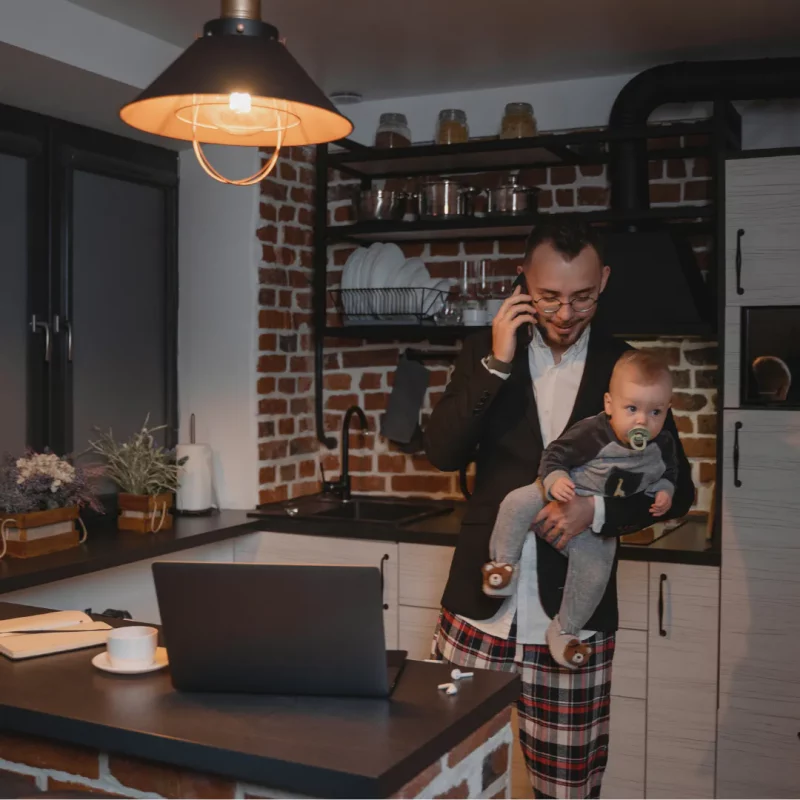 Emergency Drain Unblocking 24 Hours - Plymouth - Drain Block Services Plymouth Devon Cornwall - Drainblock Services A man in a home kitchen carries a child while talking on the phone beside an open laptop, matching a 24/7 emergency callout theme.
