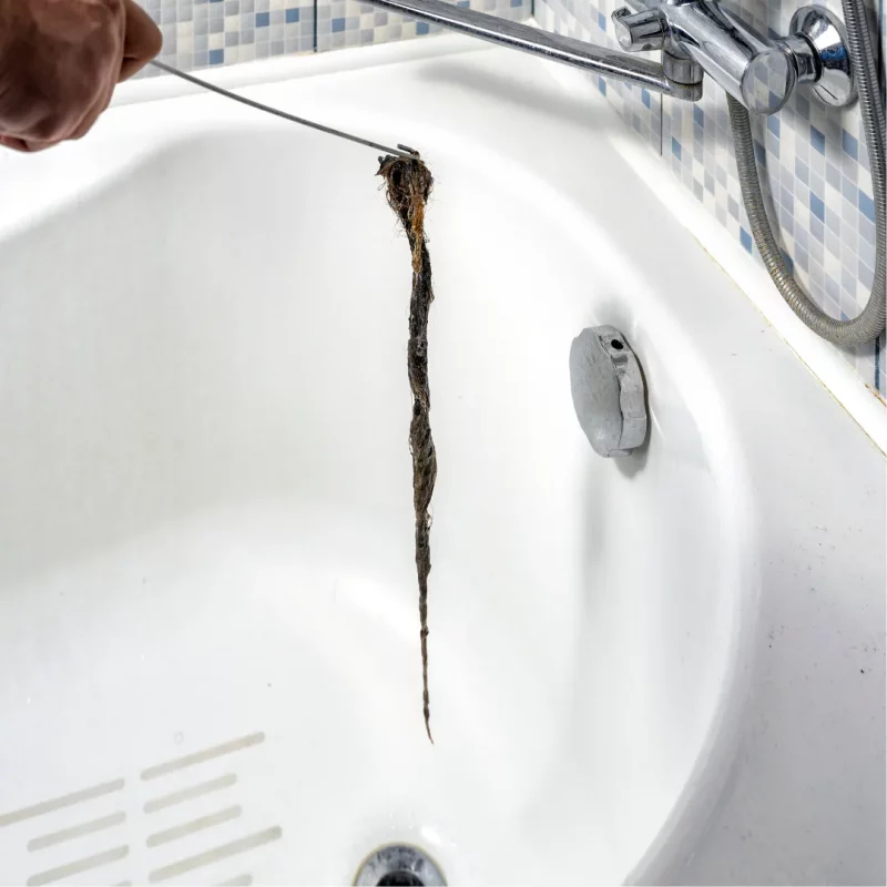 Importance of Drain Cleaning Monthly Drain Cleaning Tips Drain Block Services Plymouth Devon Cornwall - Drainblock Services Person uses a drain snake to pull a long hair clog from a bathtub drain, with tiled wall and faucet visible in the background.